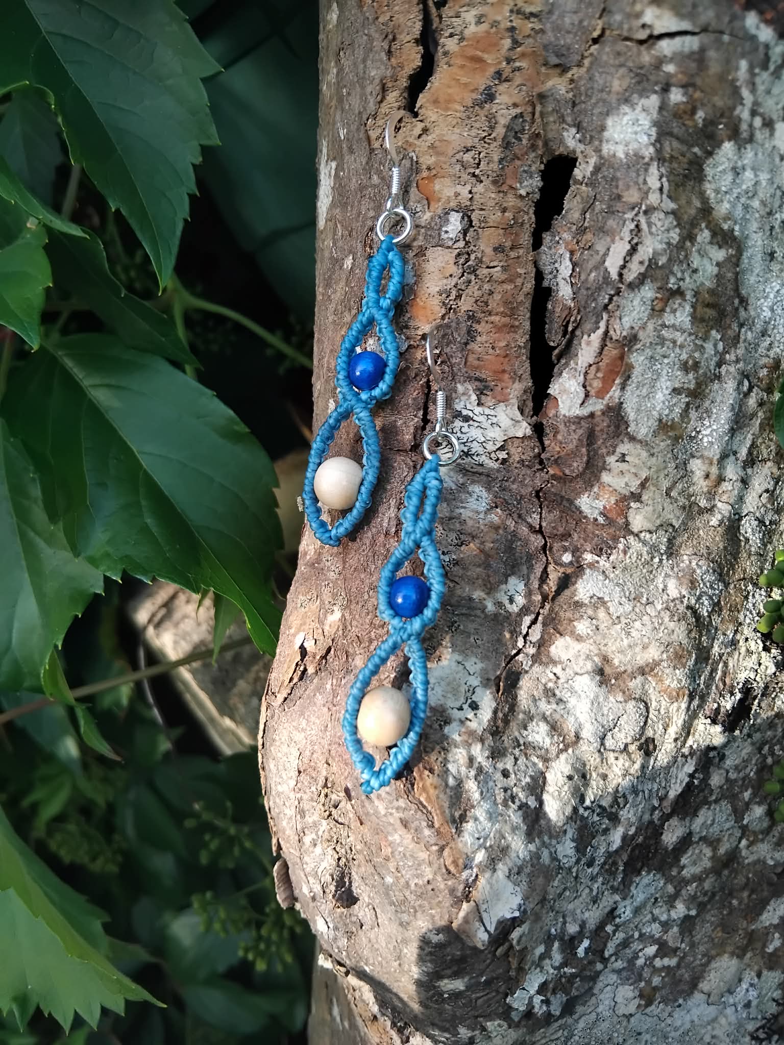 Blue and cream beaded macrame knotted earrings with silver hooks, placed on a tree branch with green leaves in the background