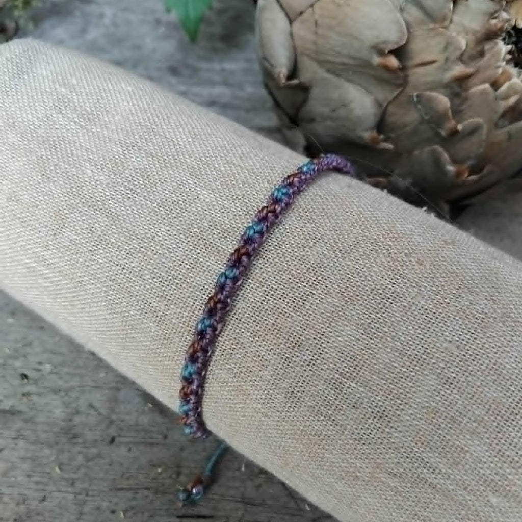 A handcrafted macrame bracelet in chocolate brown, lavender and blue toned waxed cord.  Displayed on a beige linen sleeve.