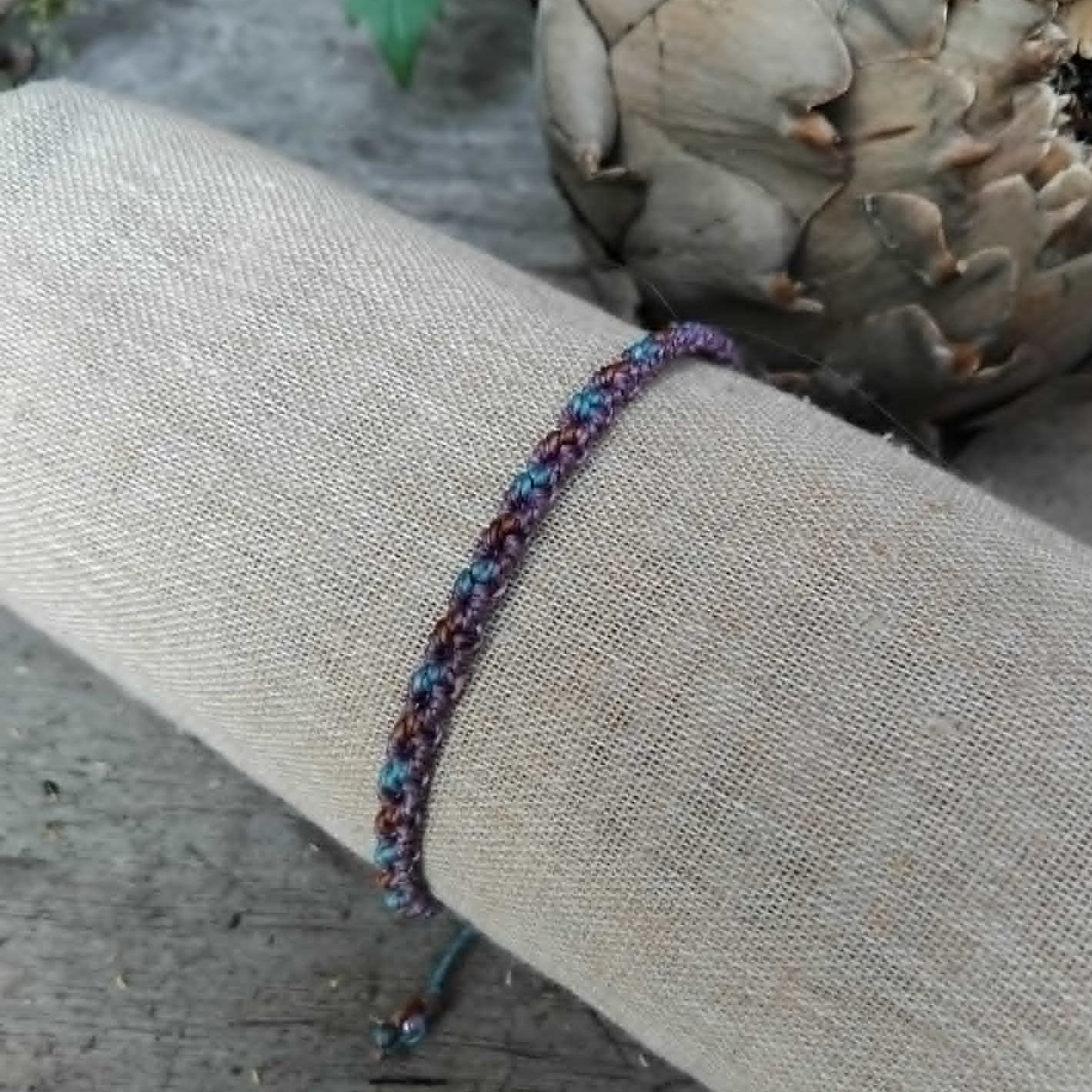 A handcrafted macrame bracelet in chocolate brown, lavender and blue toned waxed cord.  Displayed on a beige linen sleeve.