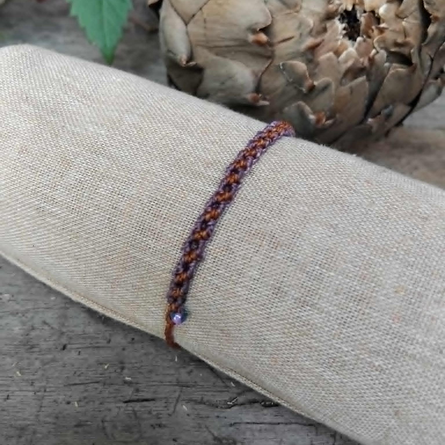 A slim macrame knotted bracelet in brown and purple tones with small lavender and haematite-toned seed beads, displayed on a rolled beige fabric sleeve against a rustic wood and artichoke background.