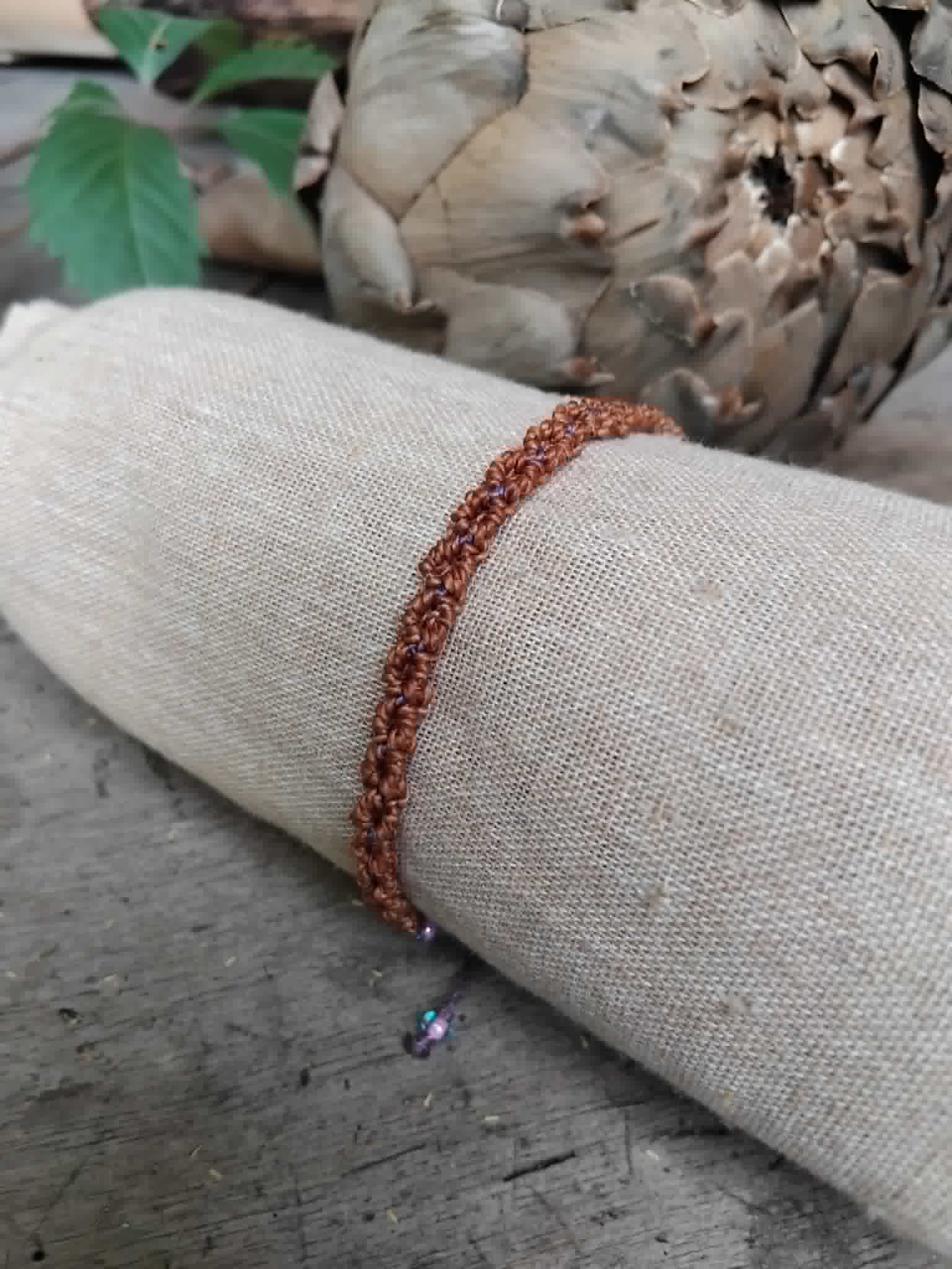 A handmade macrame bracelet in chestnut brown with lavender accents.  Displayed on a beige fabric sleeve, with a rustic background.