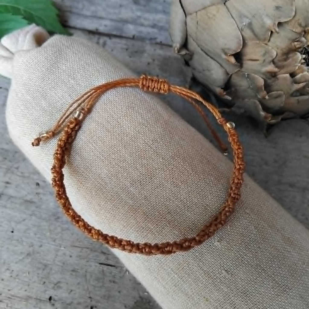 A brown macrame knotted bracelet with a small transparent and golden-toned bead accent, displayed on a beige fabric roll over a rustic gray wooden surface with decorative leaves and a dried flower in the background.