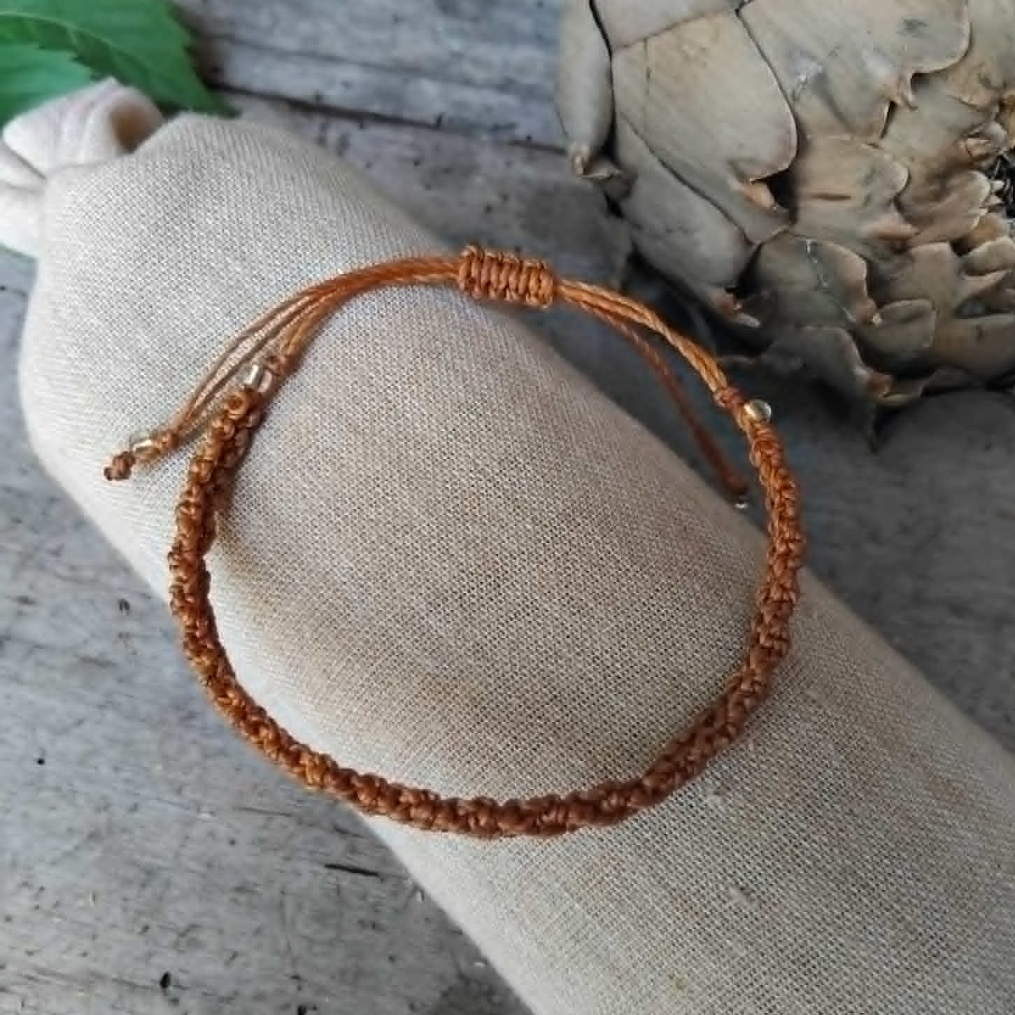 A brown macrame knotted bracelet with a small transparent and golden-toned bead accent, displayed on a beige fabric roll over a rustic gray wooden surface with decorative leaves and a dried flower in the background.