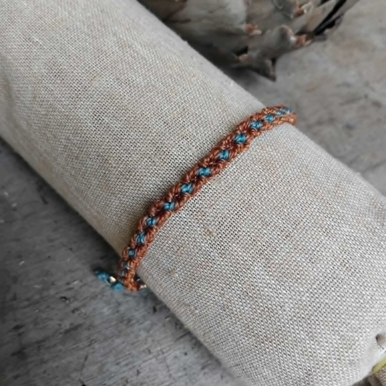 A handcrafted knotted macrame bracelet in chestnut brown with blue accents, displayed on a beige fabric sleeve.