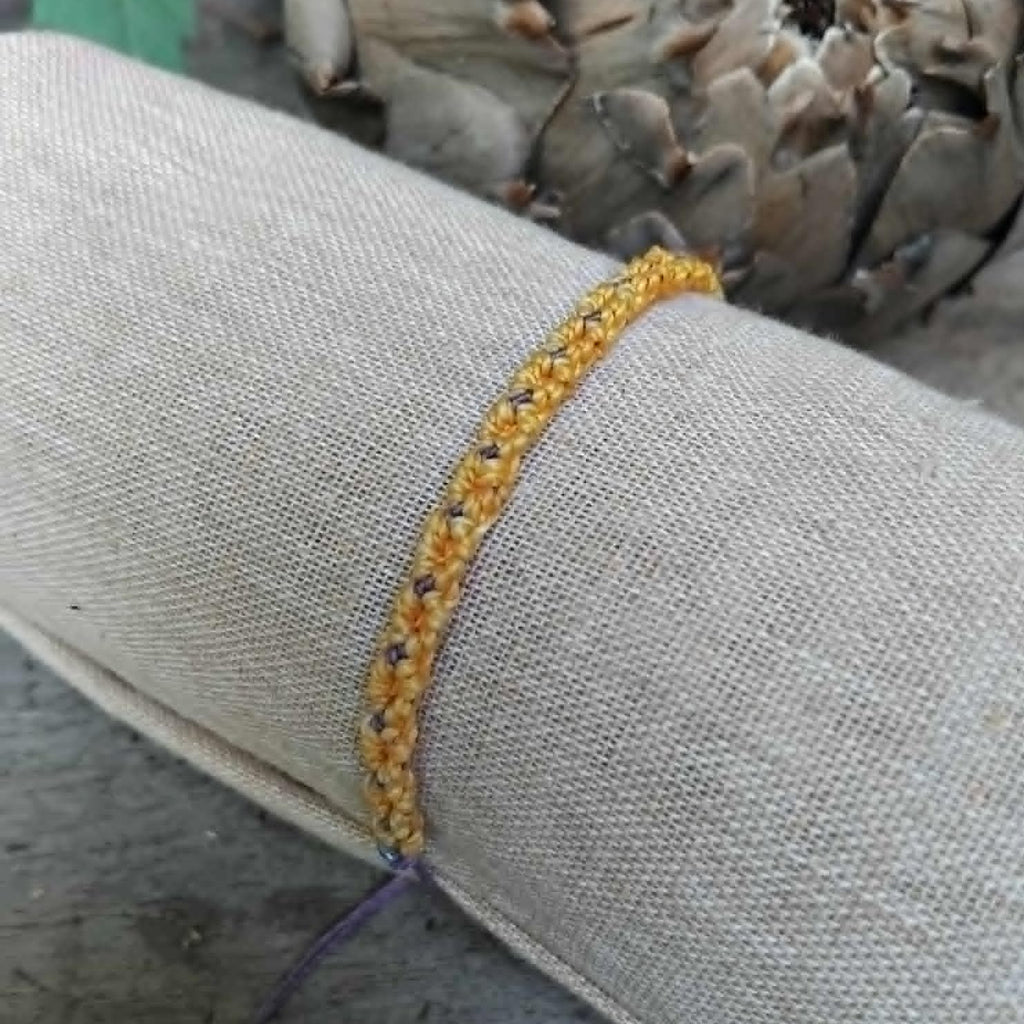 Handcrafted macrame bracelet in saffron yellow and lavender waxed cord.  Displayed on a beige linen sleeve with rustic background.
