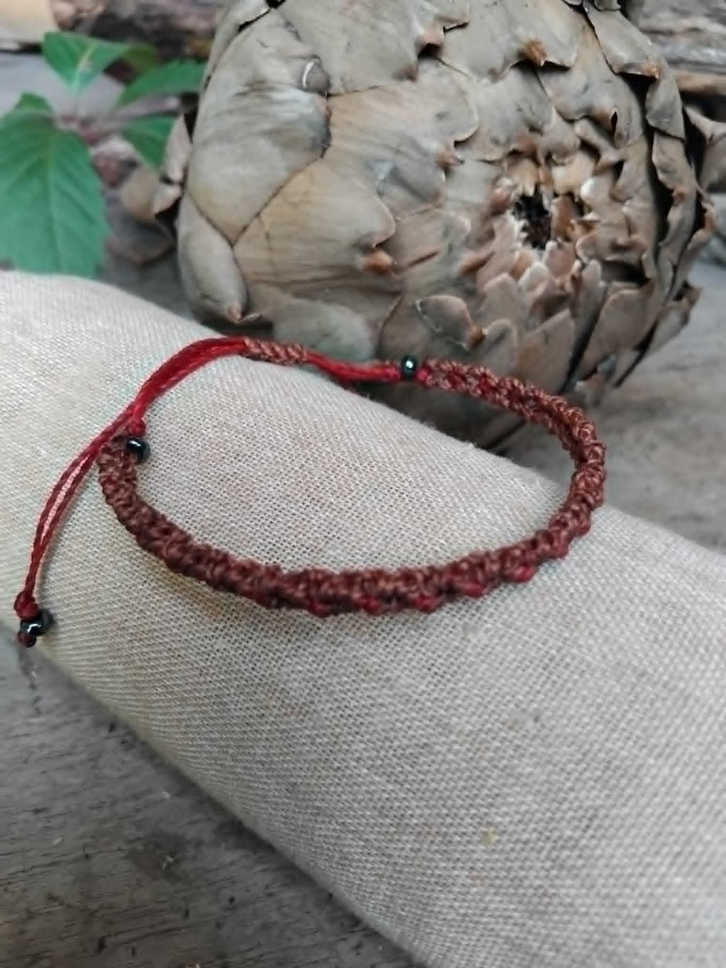 A handcrafted macrame bracelet in chocolate brown cord with burgundy red accents.  Displayed on a beige fabric sleeve, with rustic background.