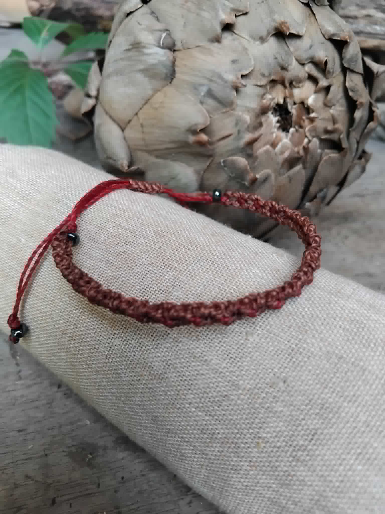 A handcrafted macrame bracelet in chocolate brown cord with burgundy red accents.  Displayed on a beige fabric sleeve, with rustic background.