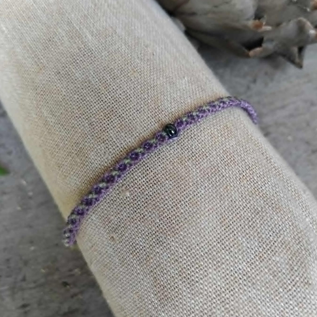 A slim macrame knotted bracelet in purple and pale lime green, featuring a small central dark bead, displayed on a beige linen fabric sleeve.