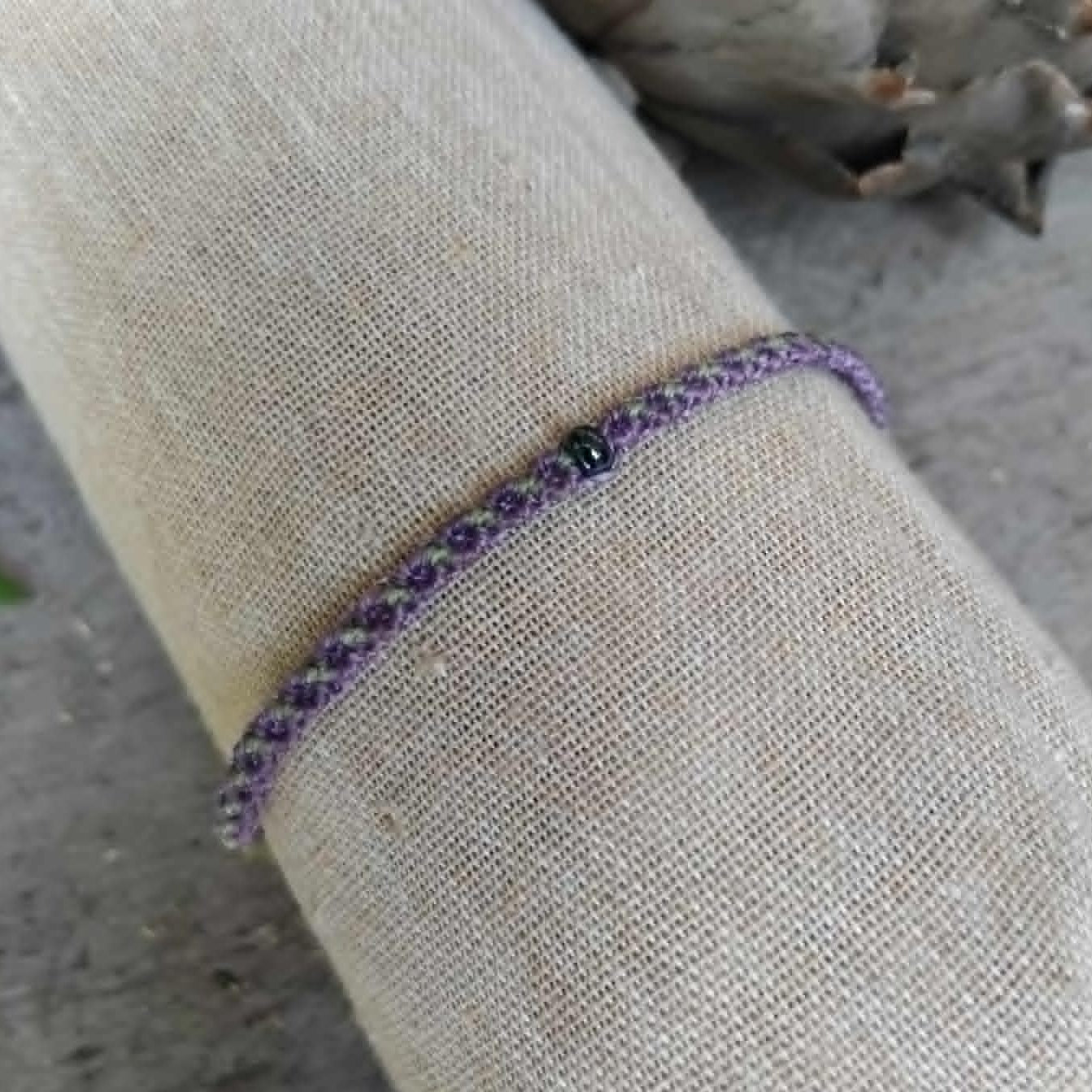 A slim macrame knotted bracelet in purple and pale lime green, featuring a small central dark bead, displayed on a beige linen fabric sleeve.