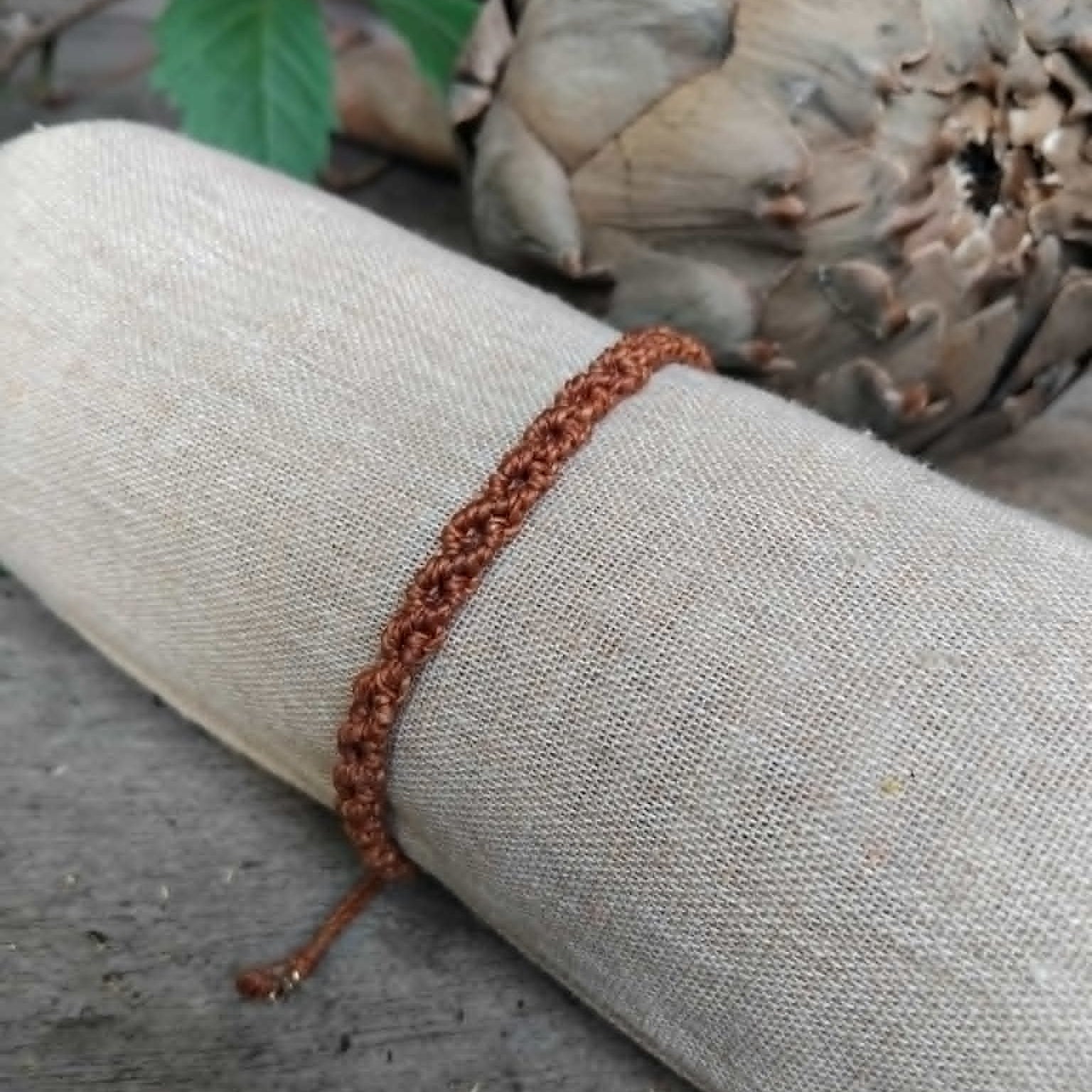 A brown macrame knotted bracelet wrapped around a rolled beige fabric sleeve, photographed on a rustic wooden surface with a dried artichoke and green leaves in the background.
