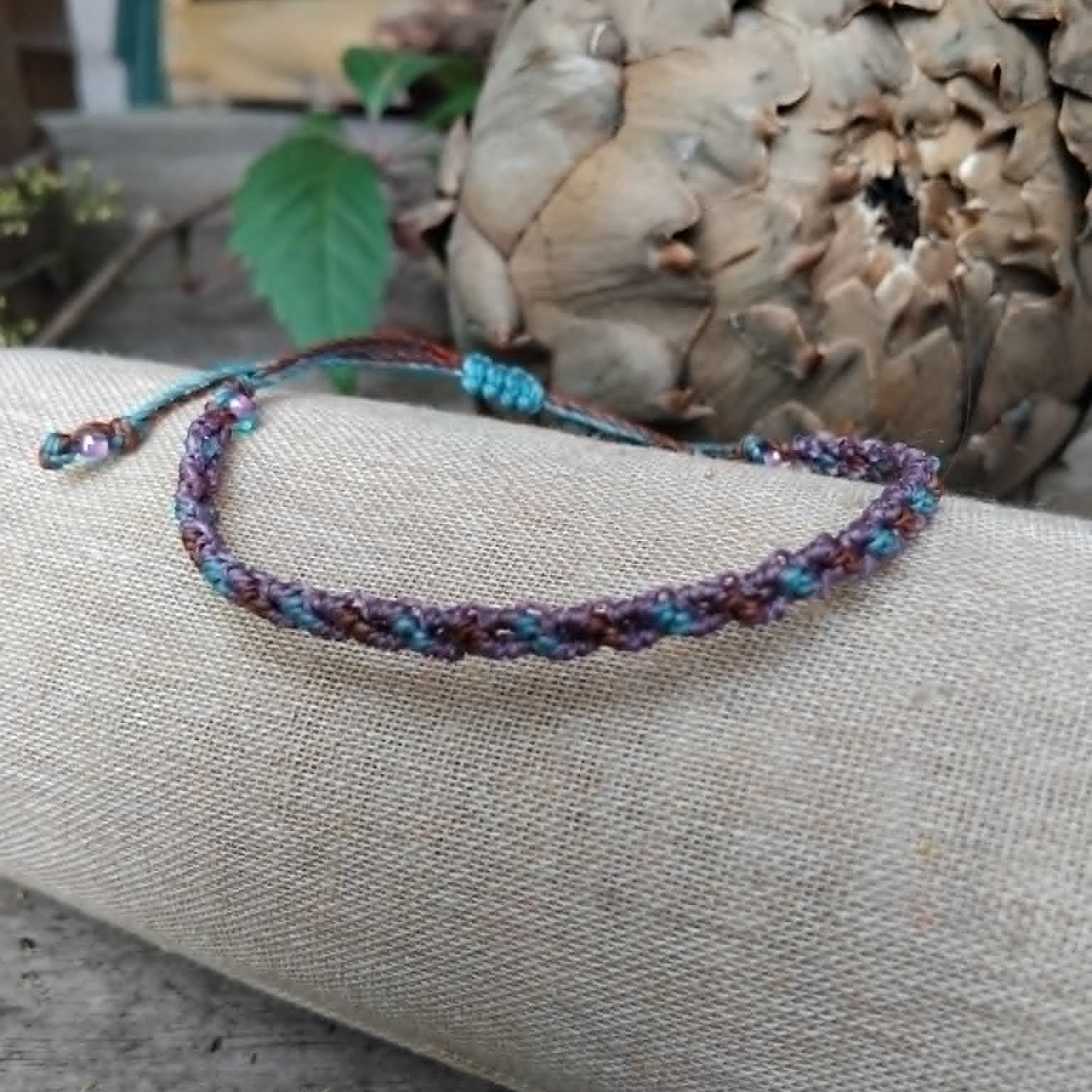 A handcrafted macrame bracelet in chocolate brown, lavender and blue toned waxed cord.  Displayed on a beige linen sleeve.