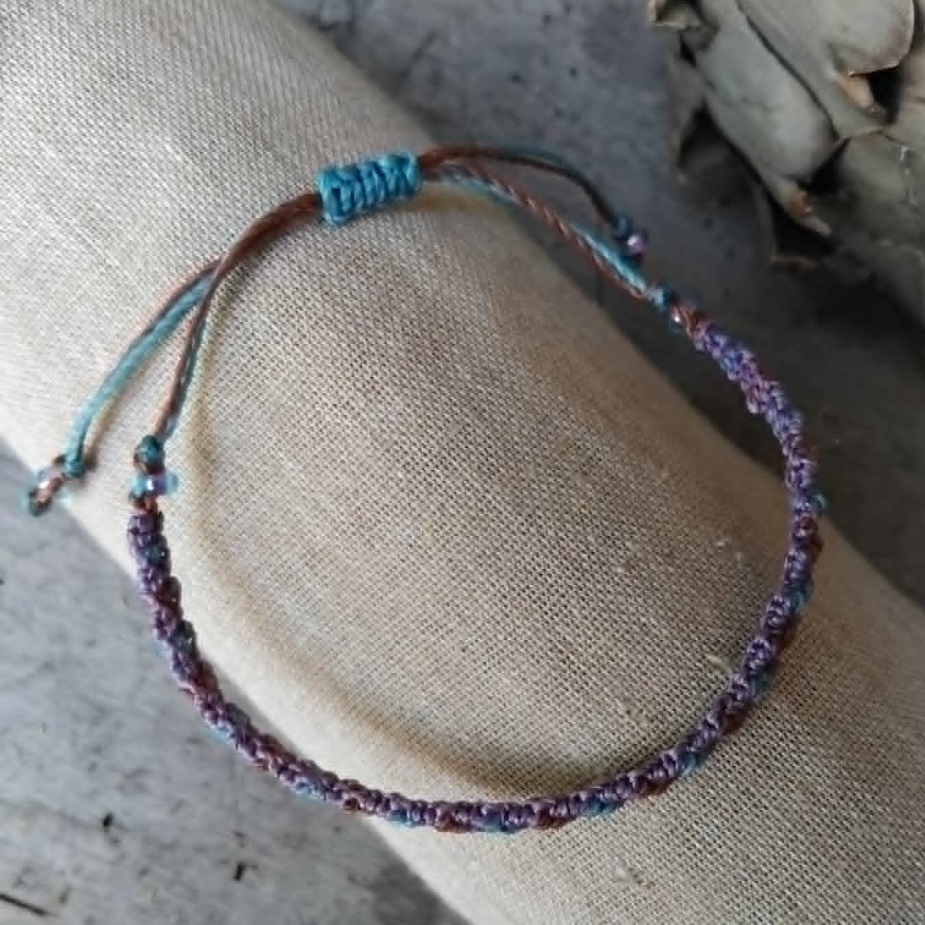 A handcrafted macrame bracelet in chocolate brown, lavender and blue toned waxed cord.  Displayed on a beige linen sleeve.