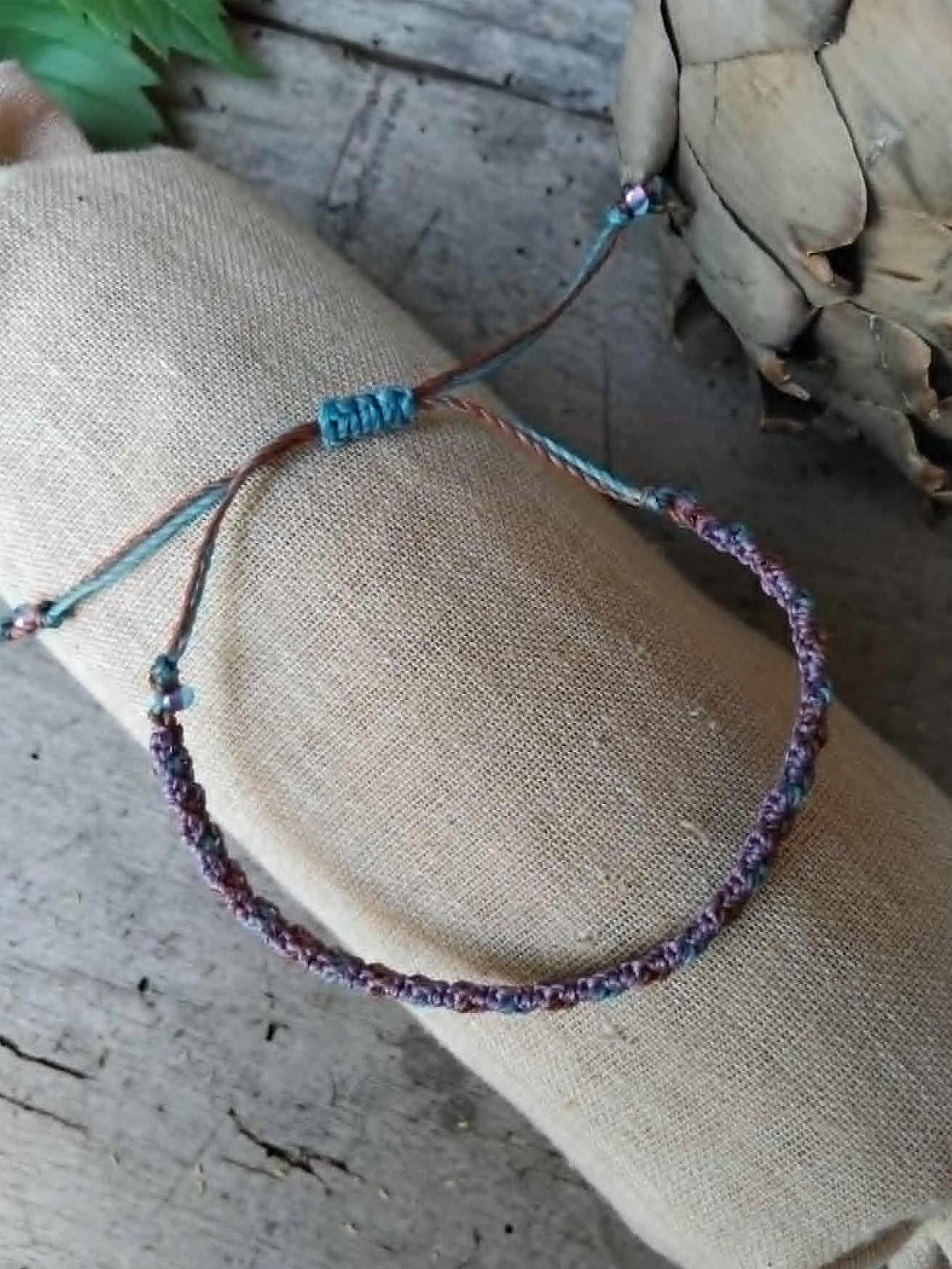 A handcrafted macrame bracelet in chocolate brown, lavender and blue toned waxed cord.  Displayed on a beige linen sleeve.