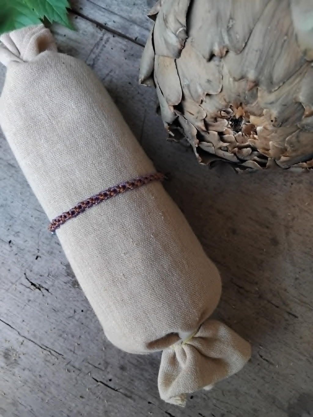 A slim macrame knotted bracelet in brown and purple tones with small lavender and haematite-toned seed beads, displayed on a rolled beige fabric sleeve against a rustic wood and artichoke background.