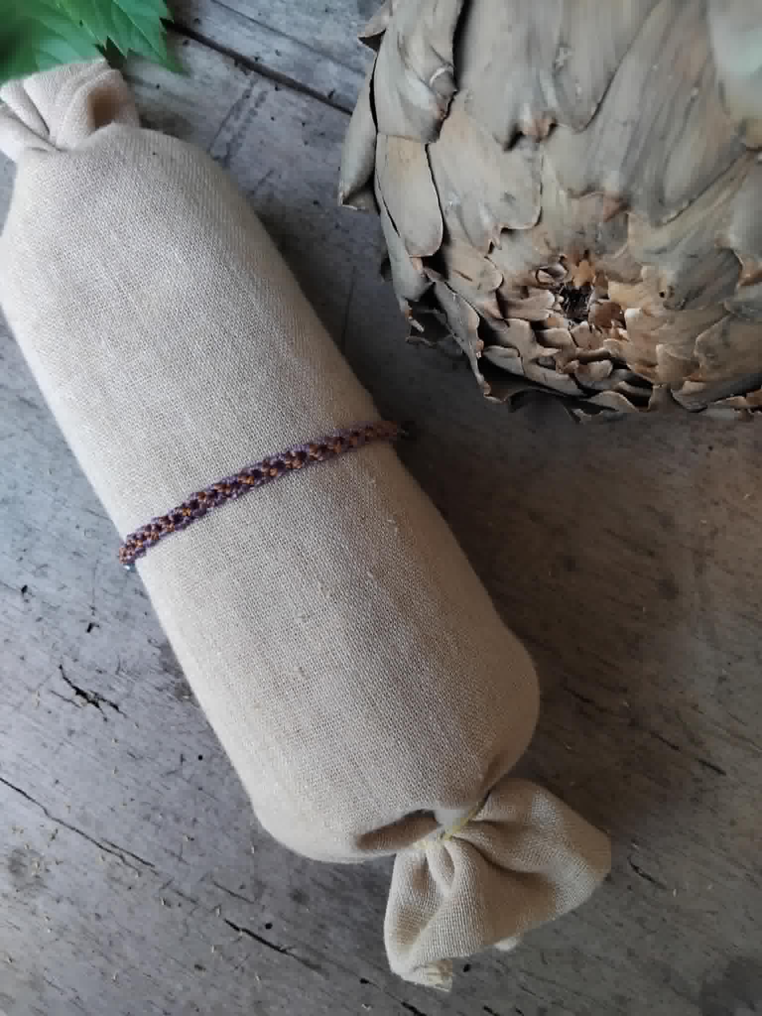 A slim macrame knotted bracelet in brown and purple tones with small lavender and haematite-toned seed beads, displayed on a rolled beige fabric sleeve against a rustic wood and artichoke background.
