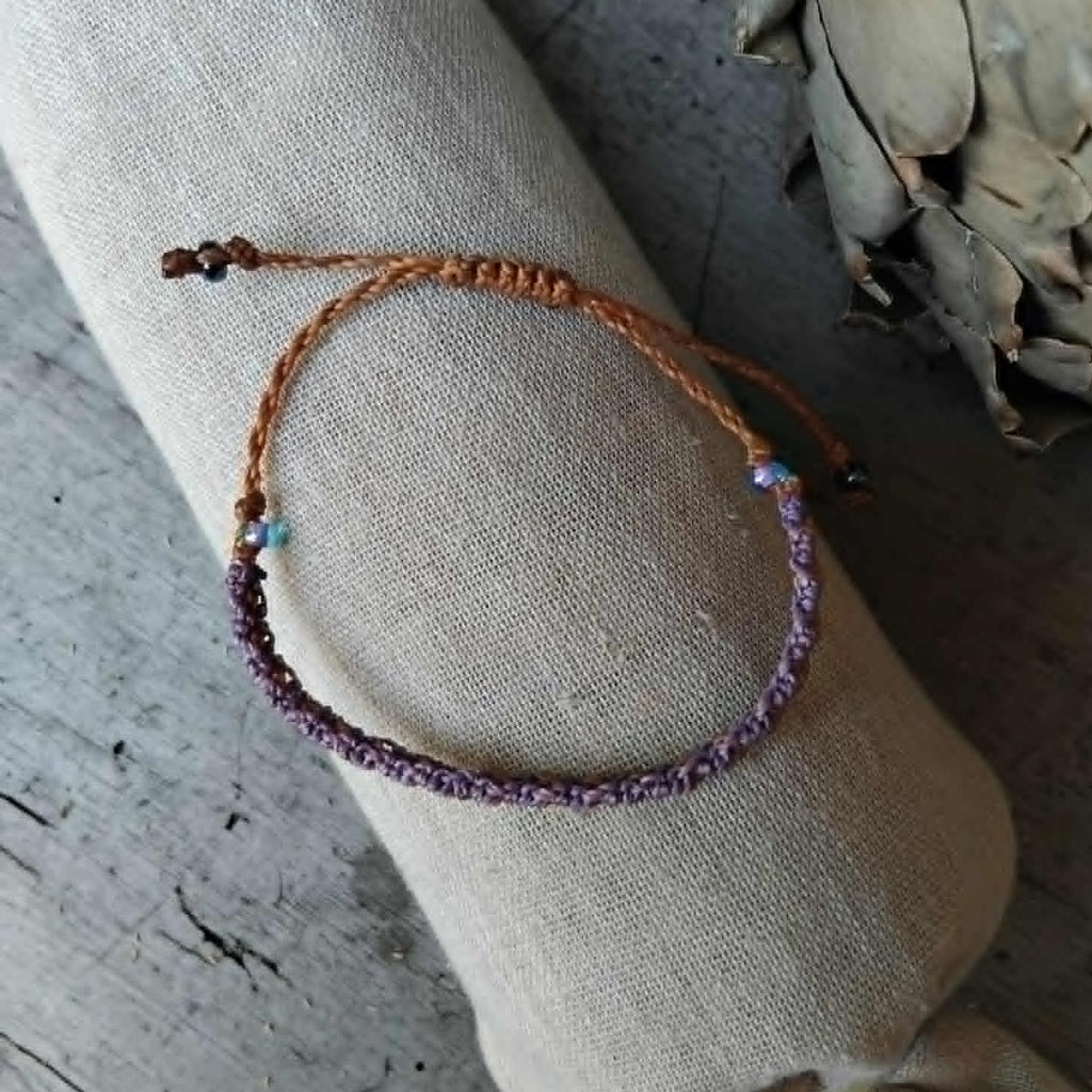 A slim macrame knotted bracelet in brown and purple tones with small lavender and haematite-toned seed beads, displayed on a rolled beige fabric sleeve against a rustic wood and artichoke background.