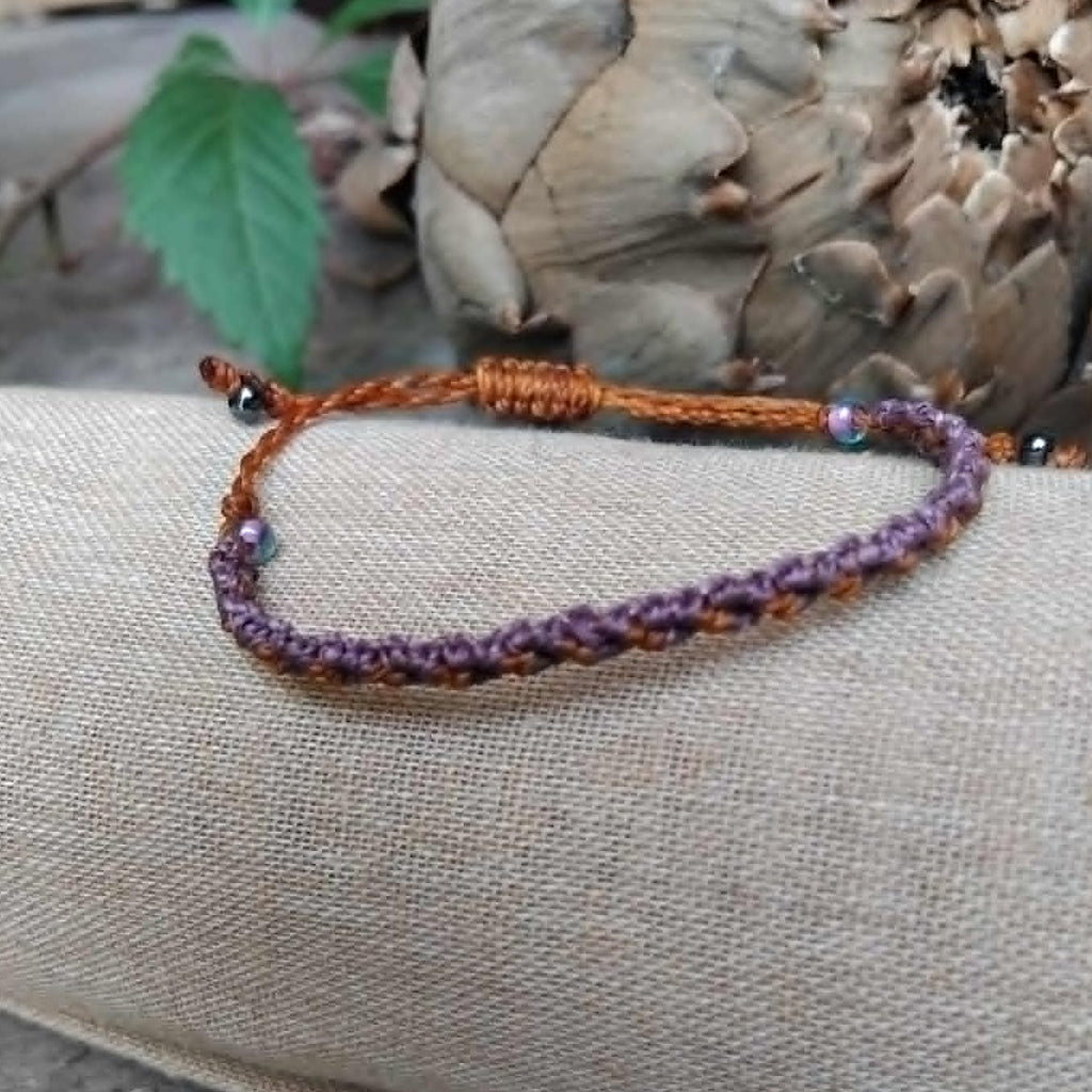 A slim macrame knotted bracelet in brown and purple tones with small lavender and haematite-toned seed beads, displayed on a rolled beige fabric sleeve against a rustic wood and artichoke background.