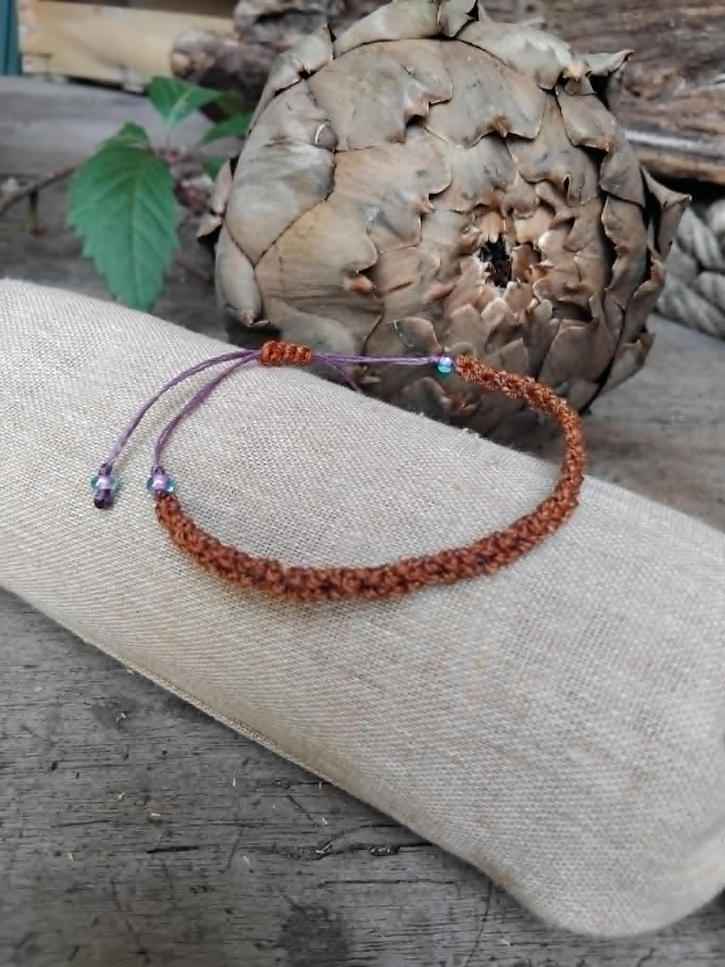 A handmade macrame bracelet in chestnut brown with lavender accents.  Displayed on a beige fabric sleeve, with a rustic background.