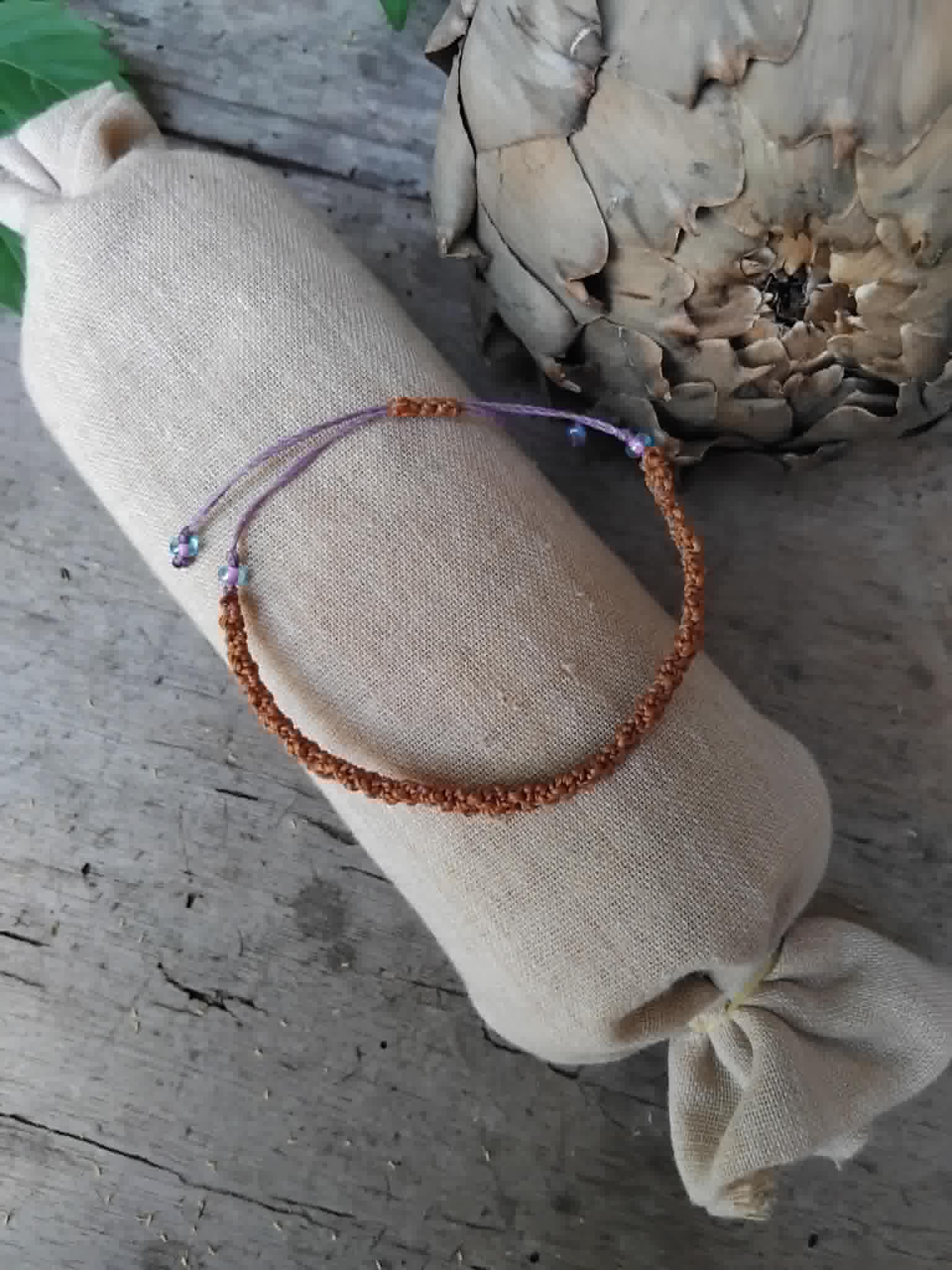 A handmade macrame bracelet in chestnut brown with lavender accents.  Displayed on a beige fabric sleeve, with a rustic background.