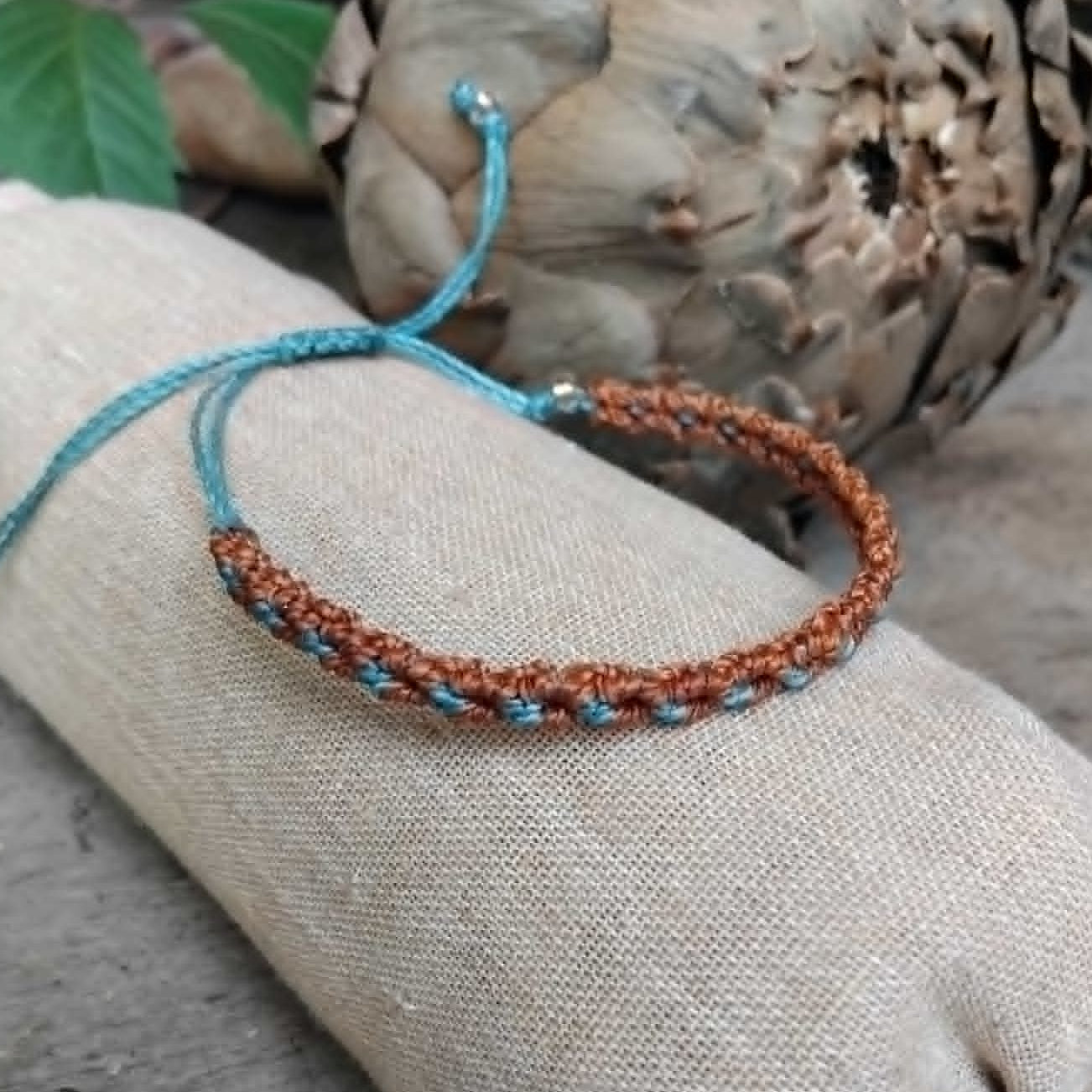 A handcrafted knotted macrame bracelet in chestnut brown with blue accents, displayed on a beige fabric sleeve.