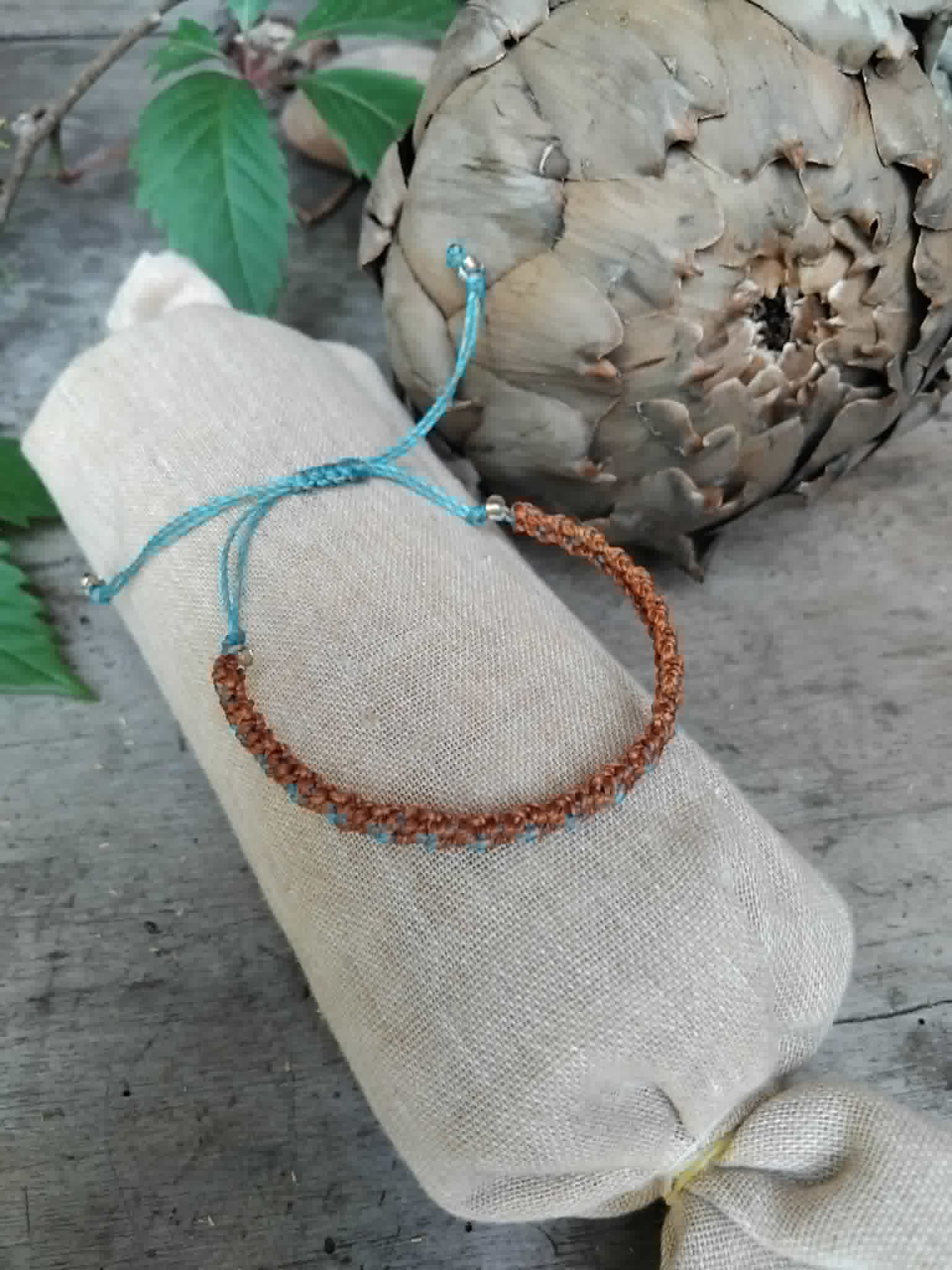 A handcrafted knotted macrame bracelet in chestnut brown with blue accents, displayed on a beige fabric sleeve.