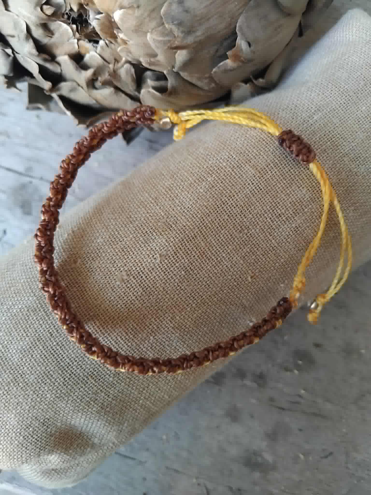 A handmade macrame bracelet in chocolate brown and saffron yellow, displayed on a beige linen fabric roll.