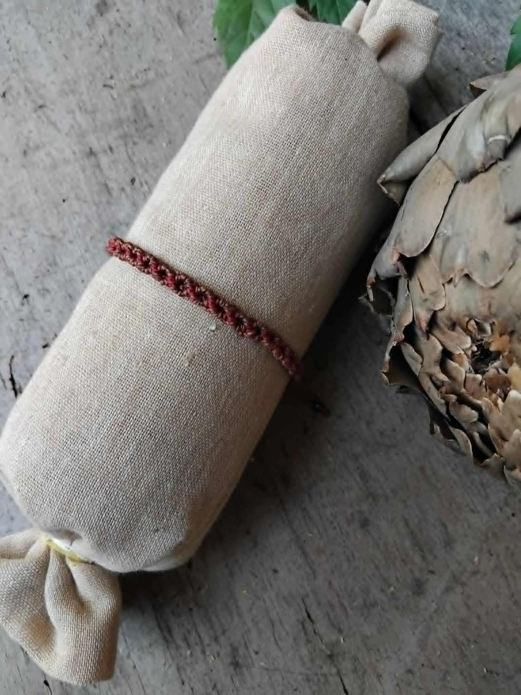 A handcrafted macrame bracelet in chocolate brown cord with burgundy red accents.  Displayed on a beige fabric sleeve, with rustic background.