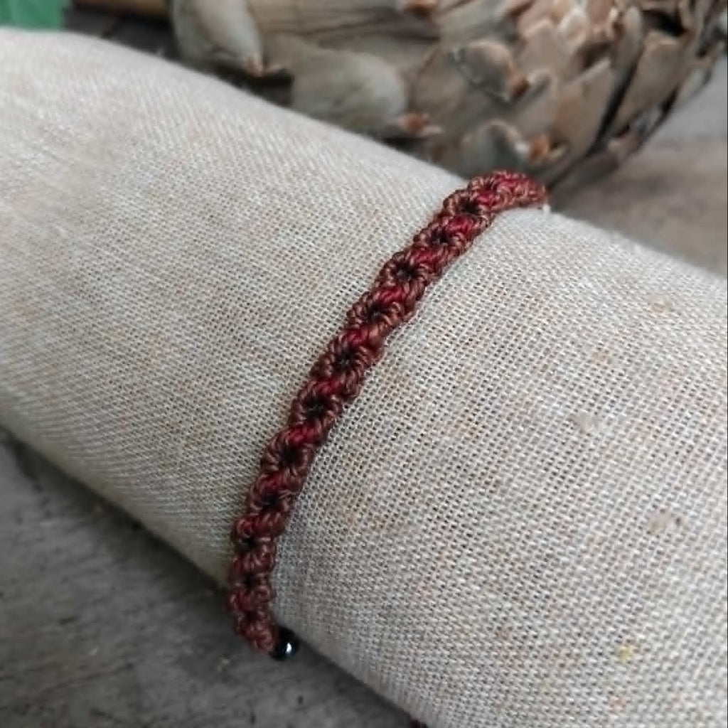 A handcrafted macrame bracelet in chocolate brown cord with burgundy red accents.  Displayed on a beige fabric sleeve, with rustic background.