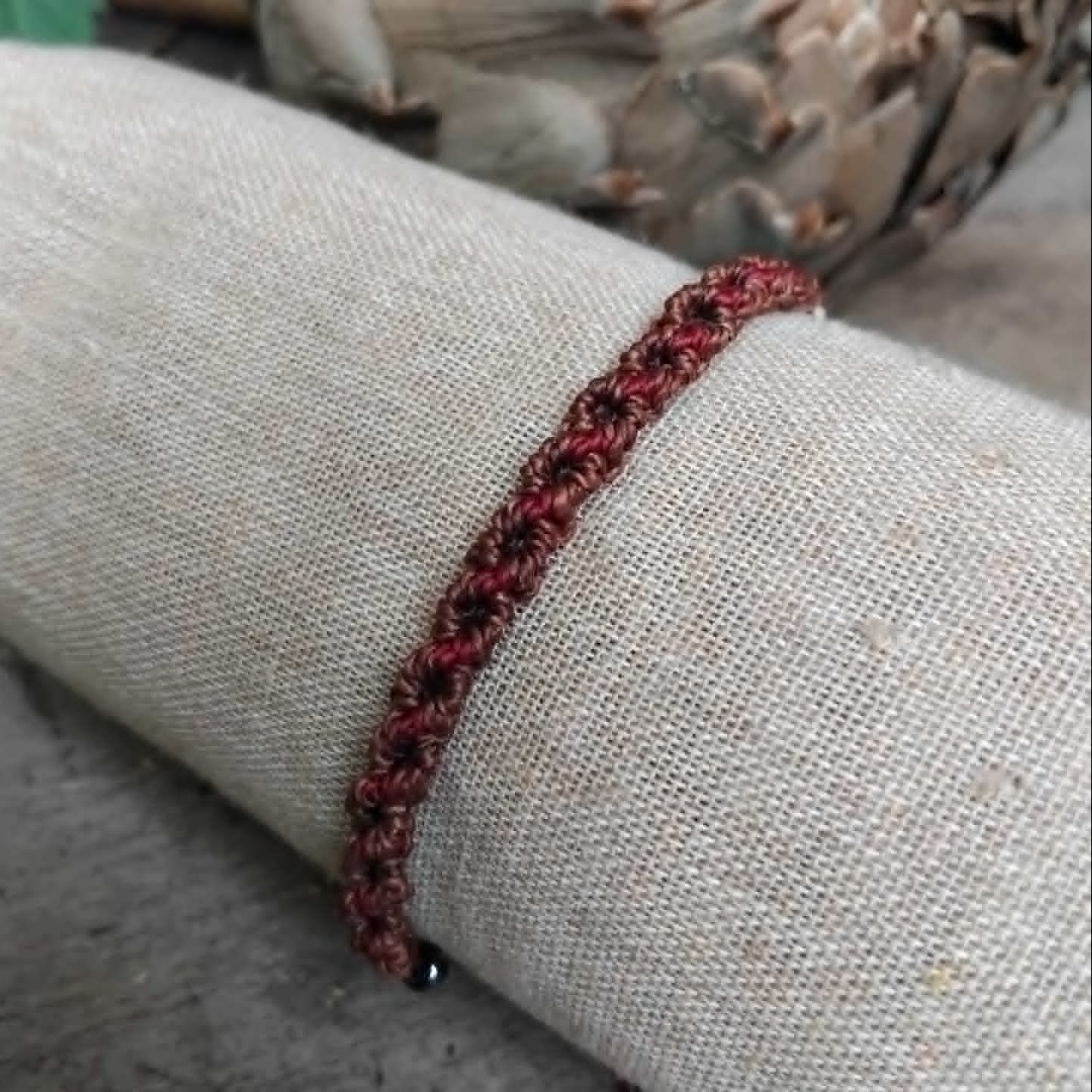 A handcrafted macrame bracelet in chocolate brown cord with burgundy red accents.  Displayed on a beige fabric sleeve, with rustic background.