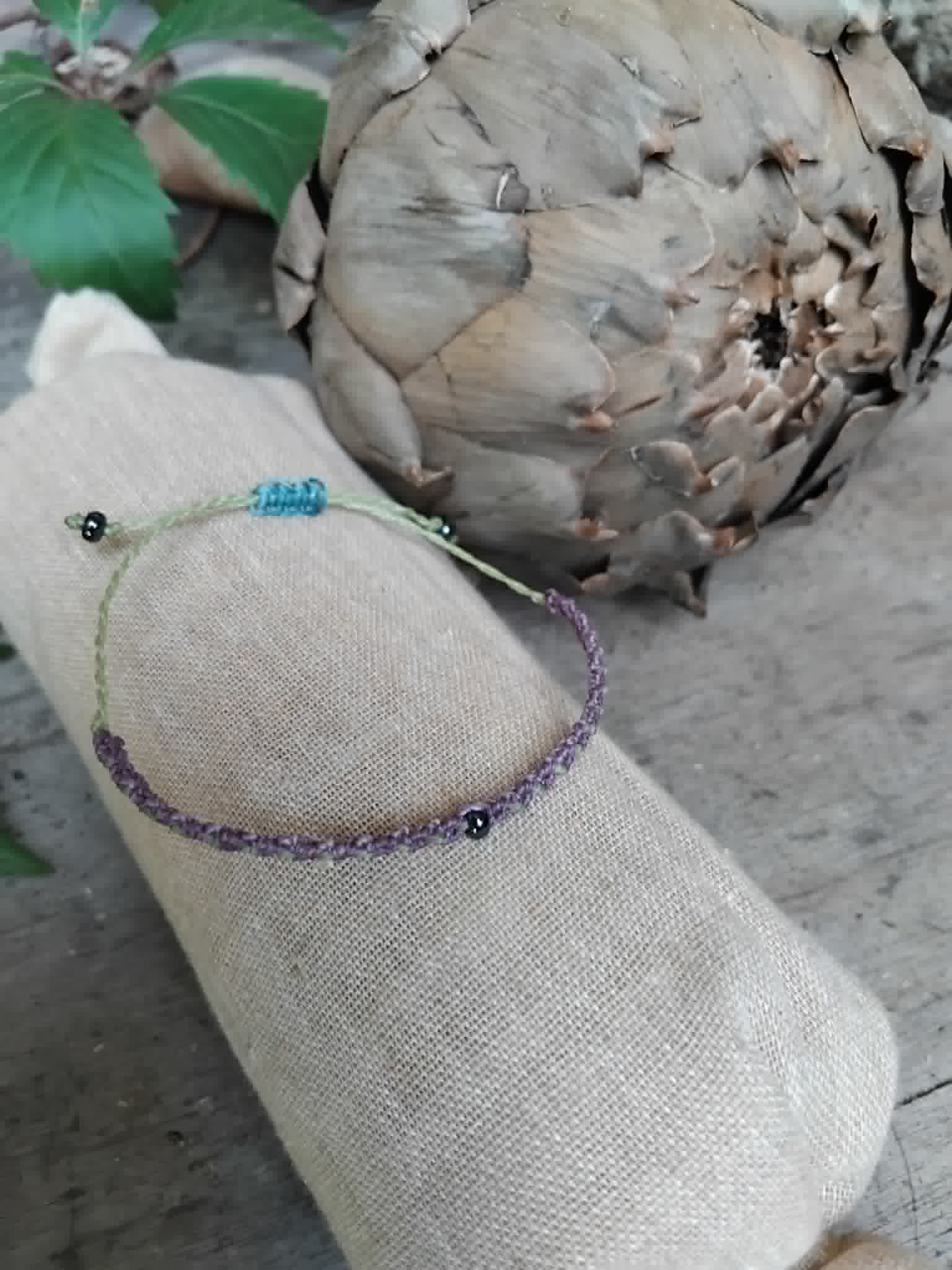 A handmade macrame bracelet in lilac and pale lime green. Has a small haematite-toned seed bead at centre and two more at each end.  Adjustable closure in blue cord.