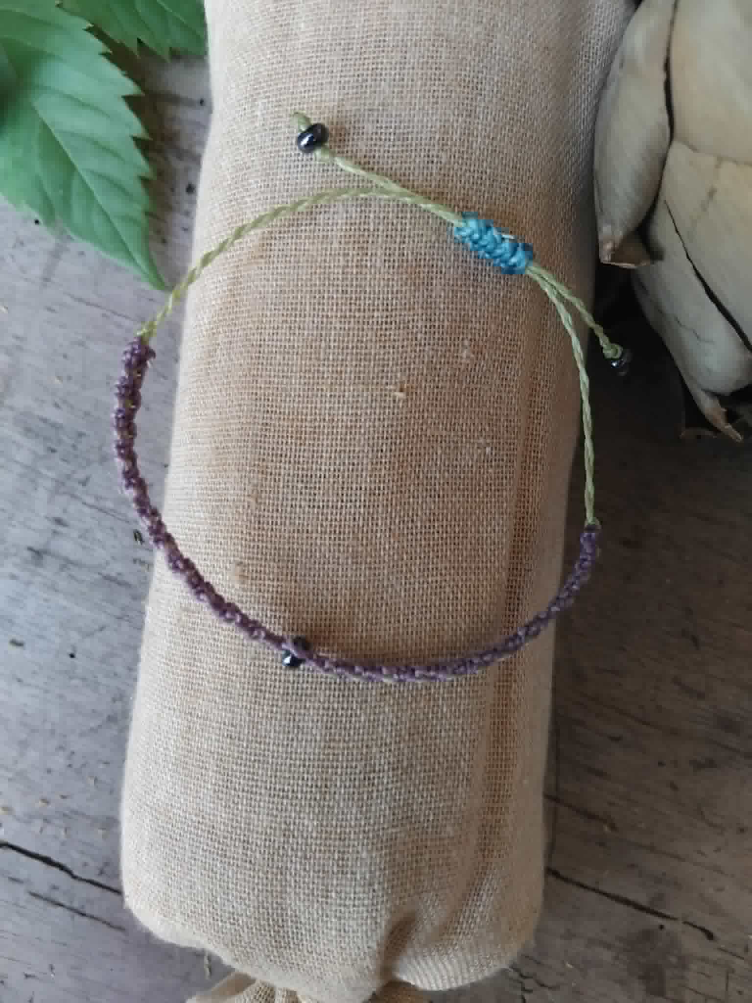 A handmade macrame bracelet in lilac and pale lime green. Has a small haematite-toned seed bead at centre and two more at each end.  Adjustable closure in blue cord.