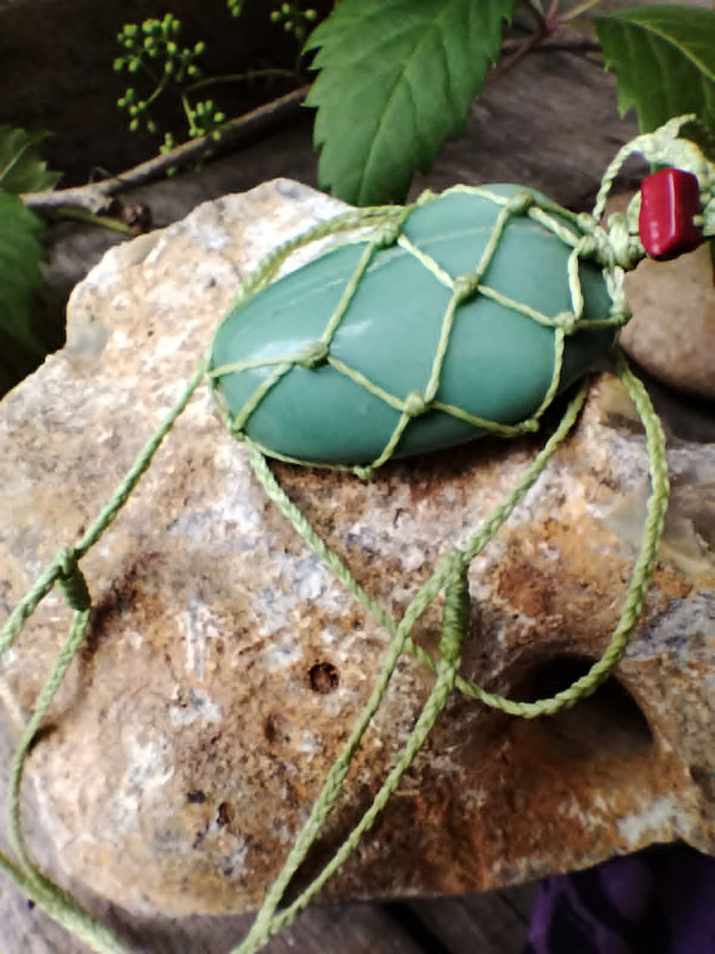 Green stone macrame knotted pendant necklace with red stone chip, and braided cord, against a rock background