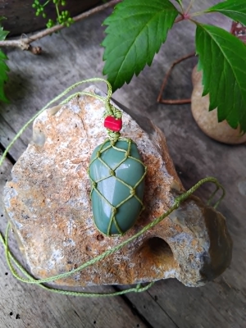 Green stone macrame pendant necklace with red stone chip and braided cord, placed on a textured rock surface.