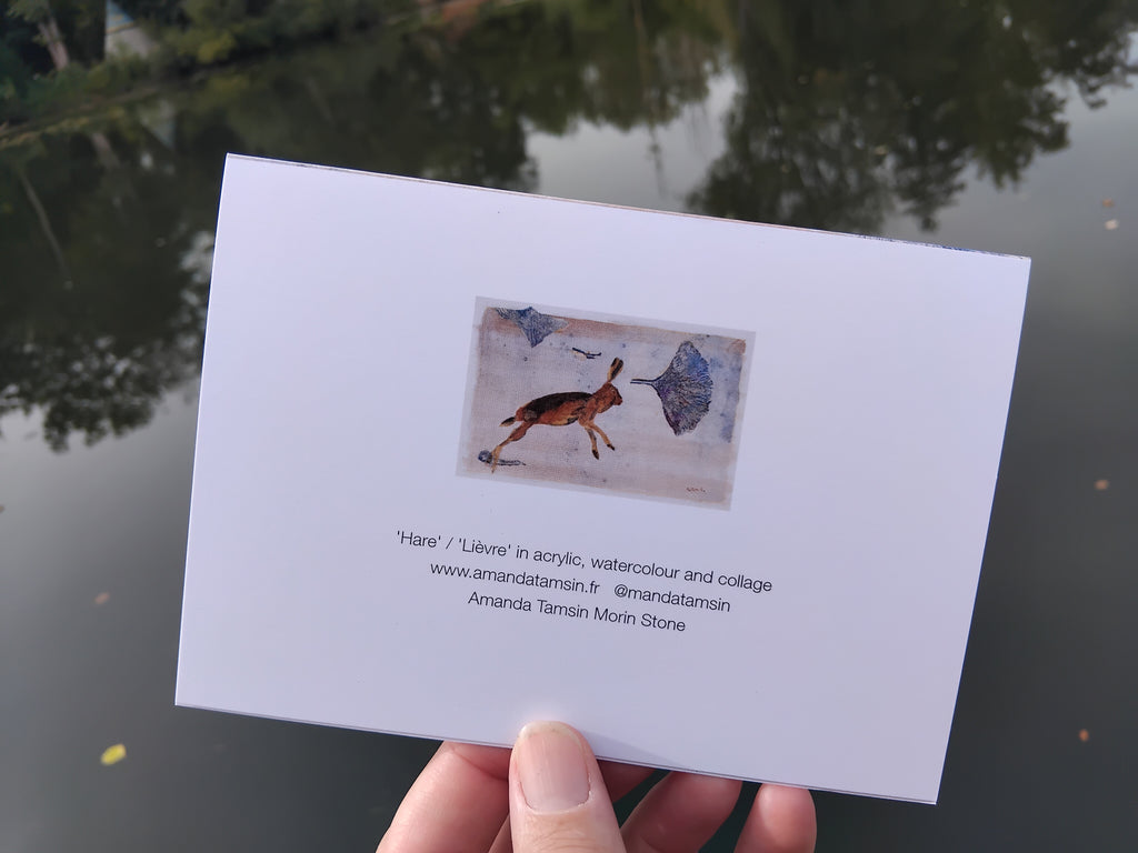 Reverse of a greetings card featuring a mini printed version of an original artwork with painted hare and ginkgo leaves, along with text about the original painting and artist.  Photographed outdoors over a reflective lake with trees in the background,