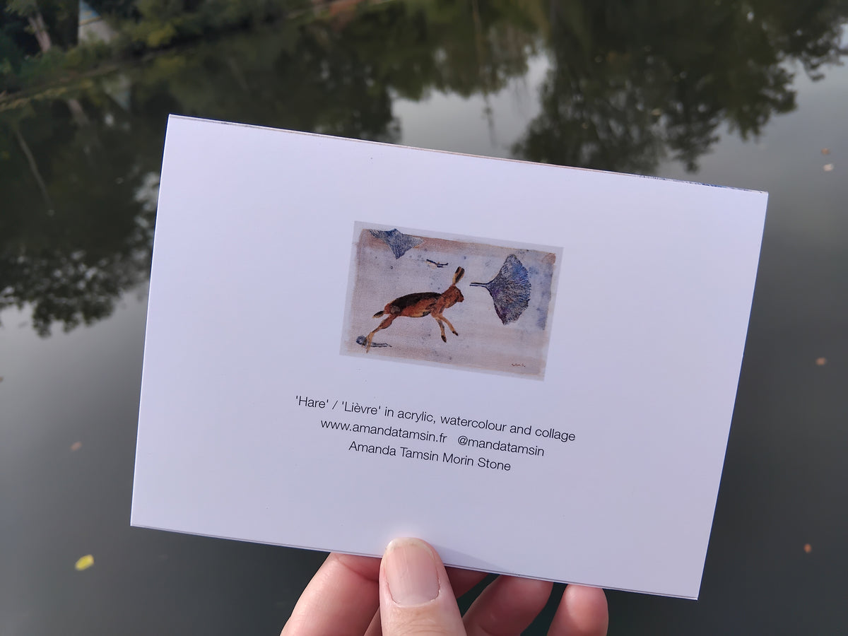 Reverse of a greetings card featuring a mini printed version of an original artwork with painted hare and ginkgo leaves, along with text about the original painting and artist.  Photographed outdoors over a reflective lake with trees in the background,