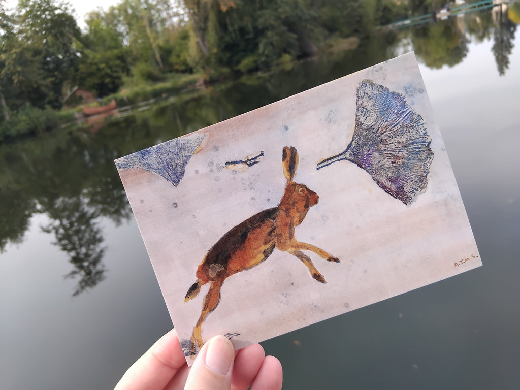 A hand holds an art Greetings Card featuring a painted hare and ginkgo leaves, photographed outdoors over a reflective lake with trees in the background.