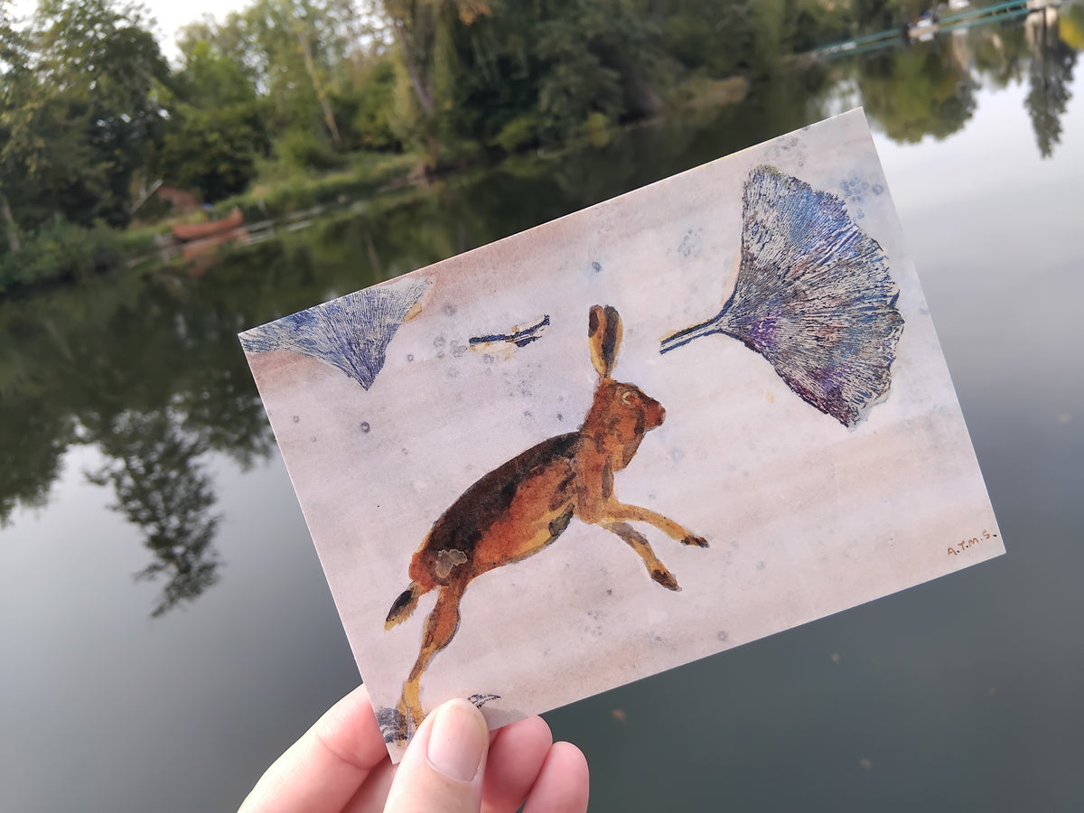 A hand holds an art Greetings Card featuring a painted hare and ginkgo leaves, photographed outdoors over a reflective lake with trees in the background.