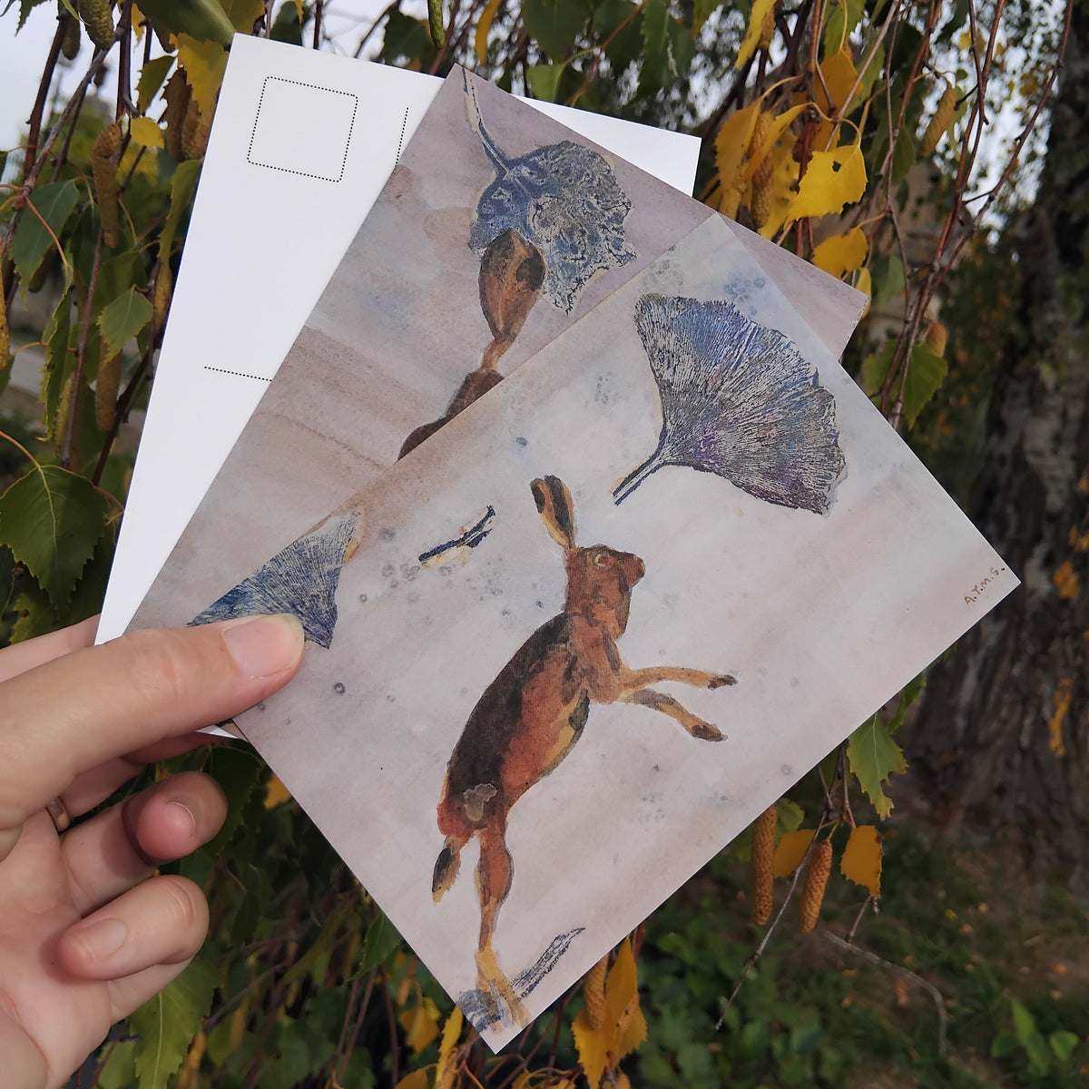 A hand holds three art postcards with painted hares and leaf print collage elements.  In earthy, indigo and blue tones.