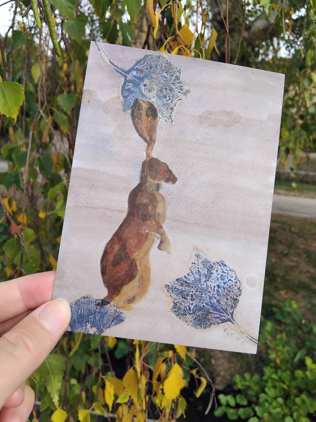 A hand holds an art postcard with painted standing hare and leaf print collage elements.  In earthy, indigo and blue tones.