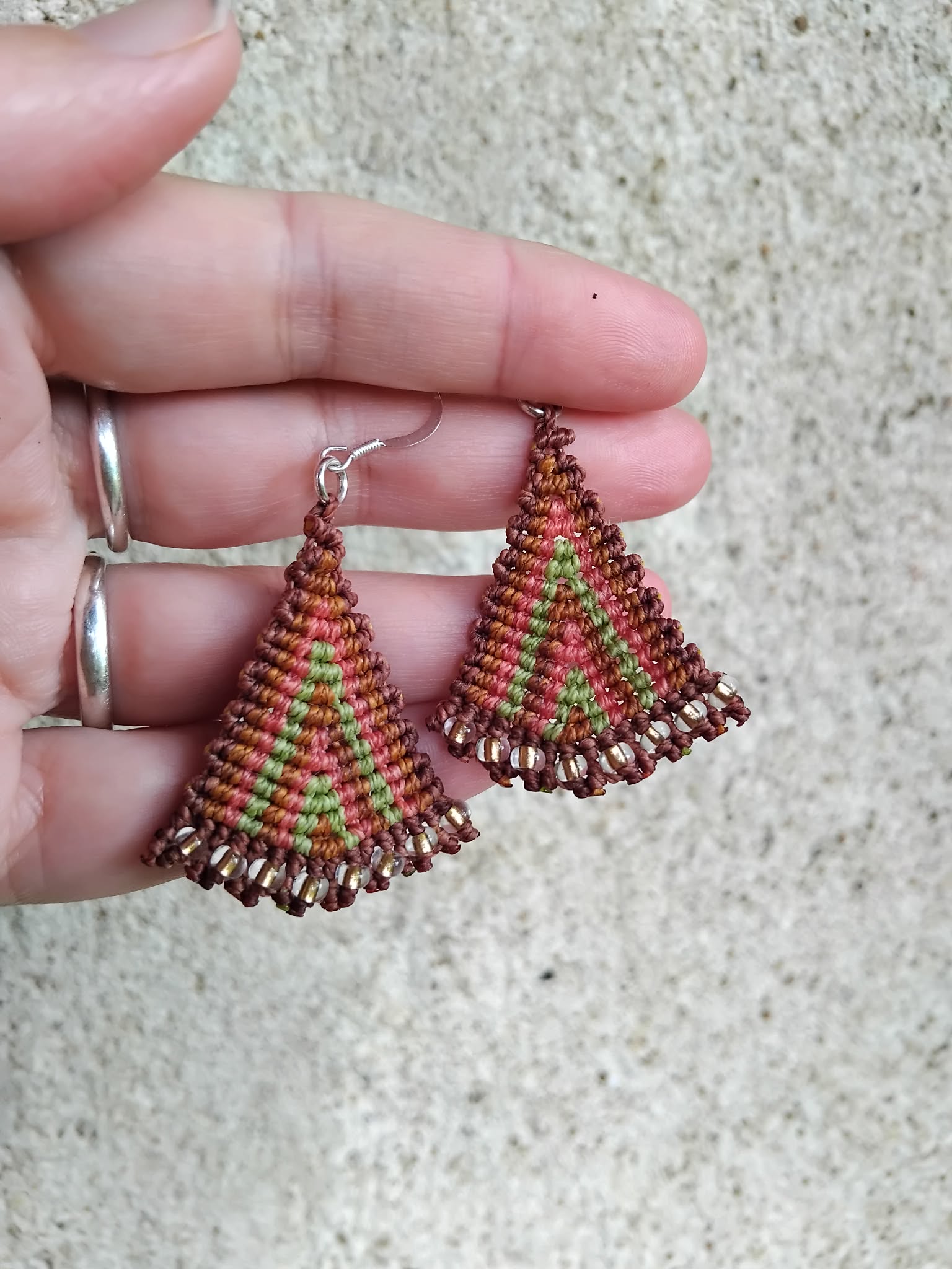 Hand holding two triangle-shaped macrame knotted and beaded earrings on a textured surface