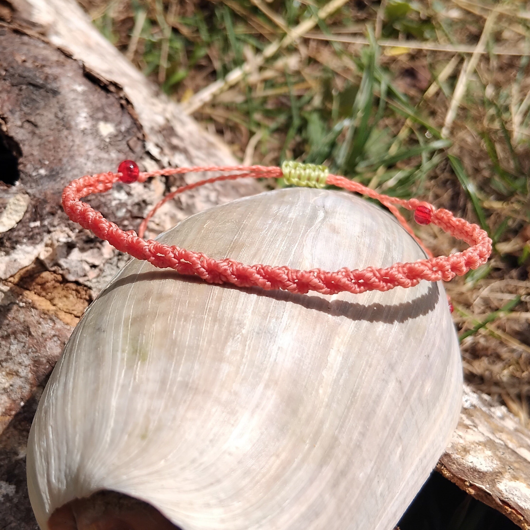 A macrame bracelet in coral pink, with a touch of apple green, placed on a shell on a wooden log outdoors