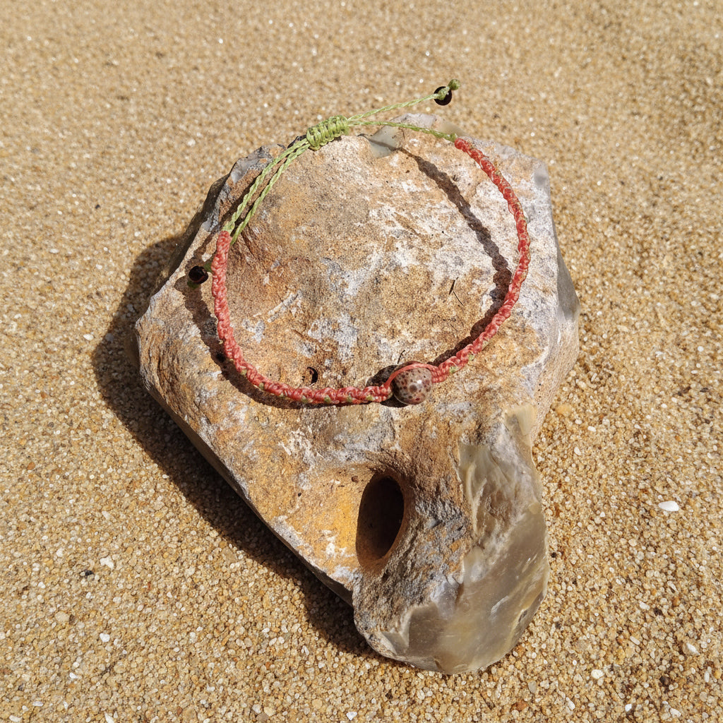 A coral and apple green toned macrame bracelet with jasper-like bead, placed on a rock, on a sandy background.